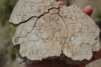 A broken piece of a home-made cement grave marker from the Varsche Fontein cemetery, held in a hand. The lettering...