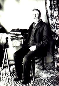 Ockert Almero Oosthuizen (1836-1922) as an old man, seated in a studio chair beside a draped table, in a dark frock...