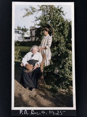 Two women in a garden. An old woman with white hair sits in a wicker chair in a white blouse and long dark skirt, a...