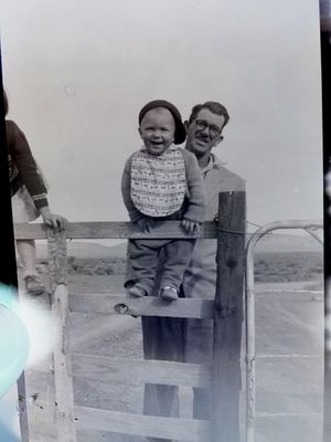 Hennie Oosthuizen at a farm gate with his son JOUBERT, about a year old, sat up on the top rail in a bib and a...