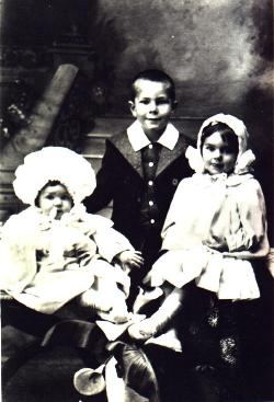 Three children in a studio, 1908: a boy standing in a buttoned jacket, a girl seated in a white bonnet and dress,...