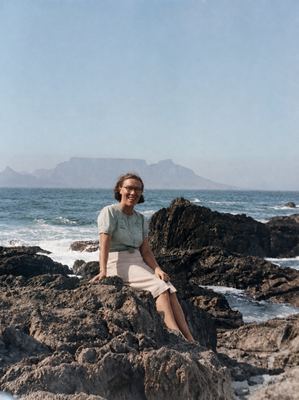 Anna Tobia Jeanne Joubert (1915-1974) sitting on the rocks at the water's edge, laughing, in a pale short-sleeved...