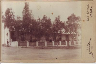 A cabinet card of a long single-storey house behind a low wall with plastered piers: a deep stoep with people...