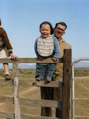 Hennie Oosthuizen at a farm gate with his son JOUBERT, about a year old, sat up on the top rail in a bib and a...