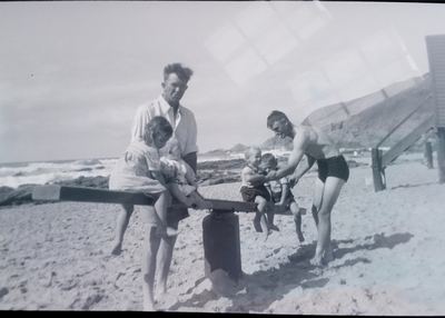 A seesaw on the sand at Herolds Bay, a plank over a drum. A girl leans on the low end holding a baby; two small boys...