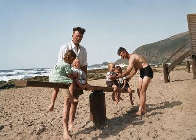 A seesaw on the sand at Herolds Bay, a plank over a drum. A girl leans on the low end holding a baby; two small boys...