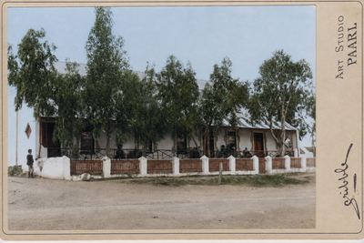 A cabinet card of a long single-storey house behind a low wall with plastered piers: a deep stoep with people...